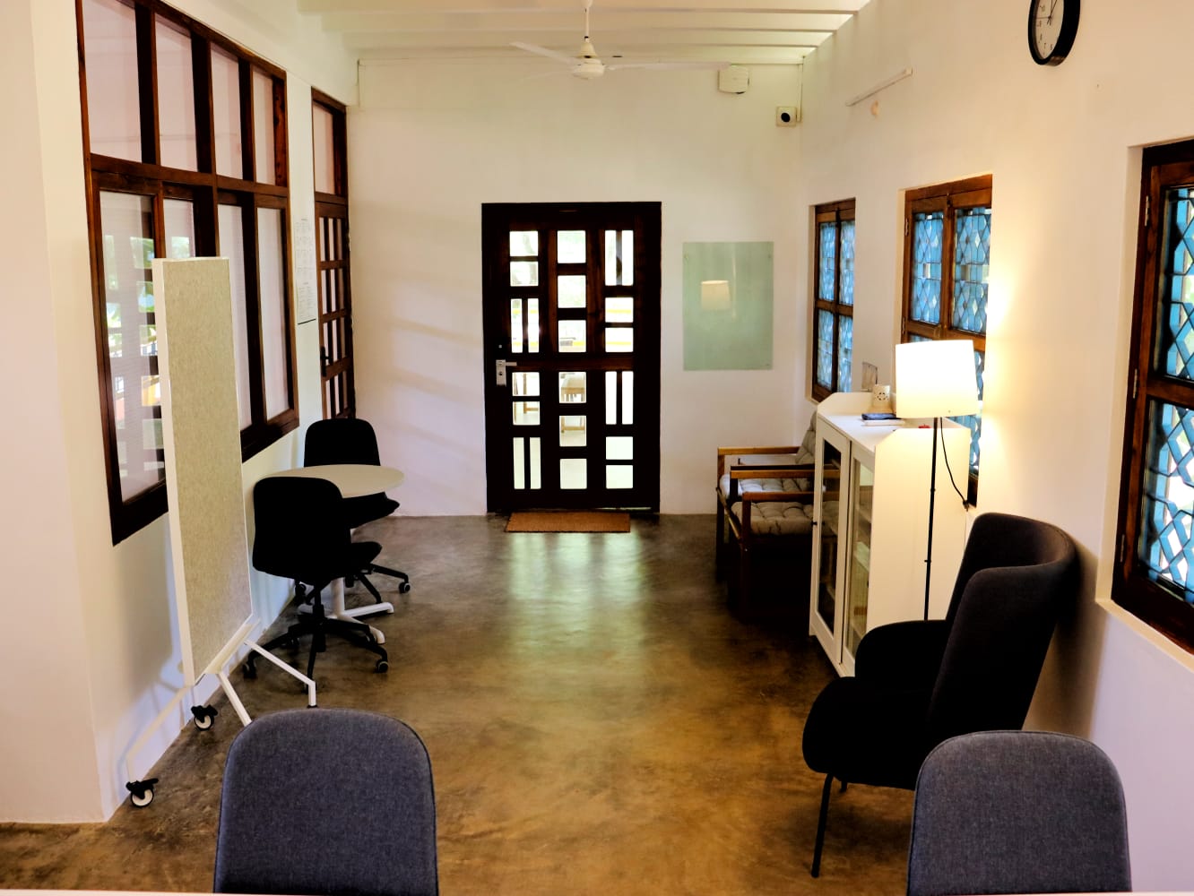 The main room looking towards a tall teak entrance door, with task chairs, a mobile pinboard and armchairs along the wall.